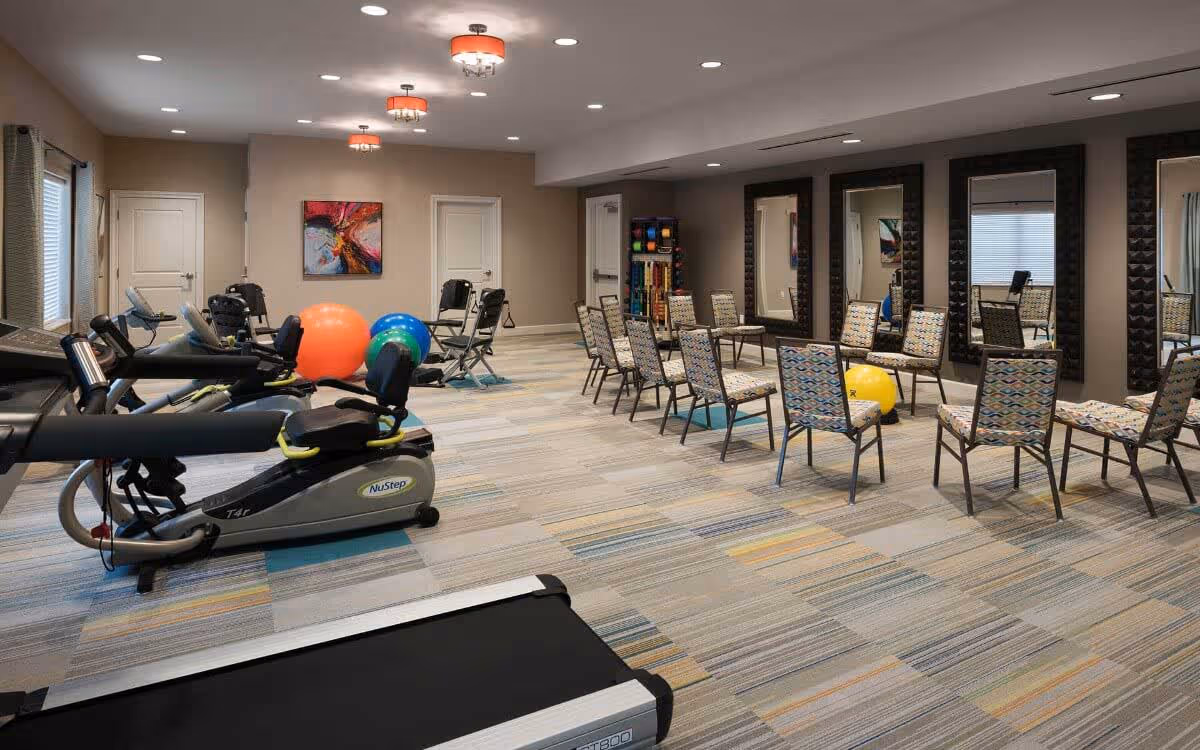 An interior fitness/therapy room with cardio machines, exercise balls, rows of chairs and large mirrors.