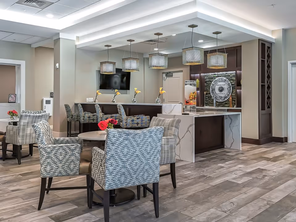 A modern senior living common area with a seating arrangement including patterned armchairs around a round table with a small flower vase. In the background, there is a bar counter with high chairs and pendant lights hanging from the ceiling. The area has a light and neutral color palette with wood-like flooring and a TV mounted on the wall.