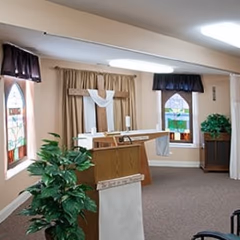 Inside a small chapel with a wooden altar, lectern, stained glass windows, and plants.