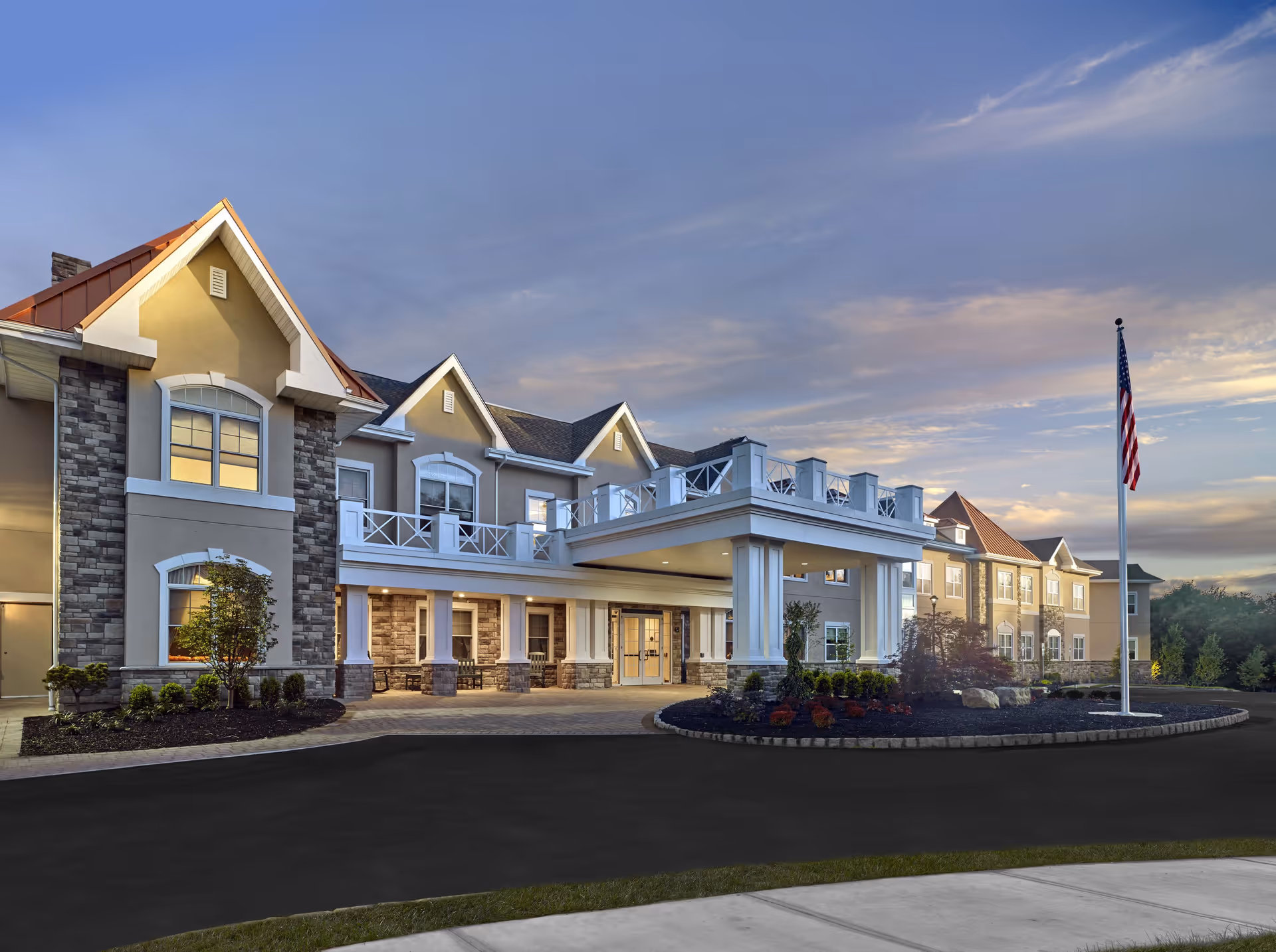 Exterior front entrance of a multi-story senior living building with columns, balconies, landscaped driveway and an American flag at dusk.