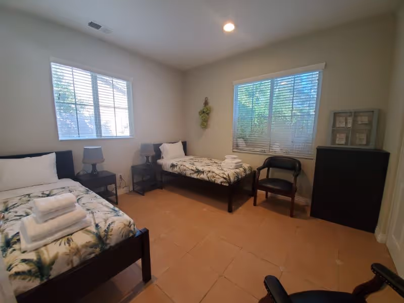 A simple bedroom with two single beds, each with a pillow and folded towels on top. The beds have matching floral bedspreads. There are two windows with blinds letting in natural light. Between the beds are two small nightstands with lamps. A black chair and a black dresser with framed pictures on top are also visible. The floor is tiled and the walls are painted light beige.