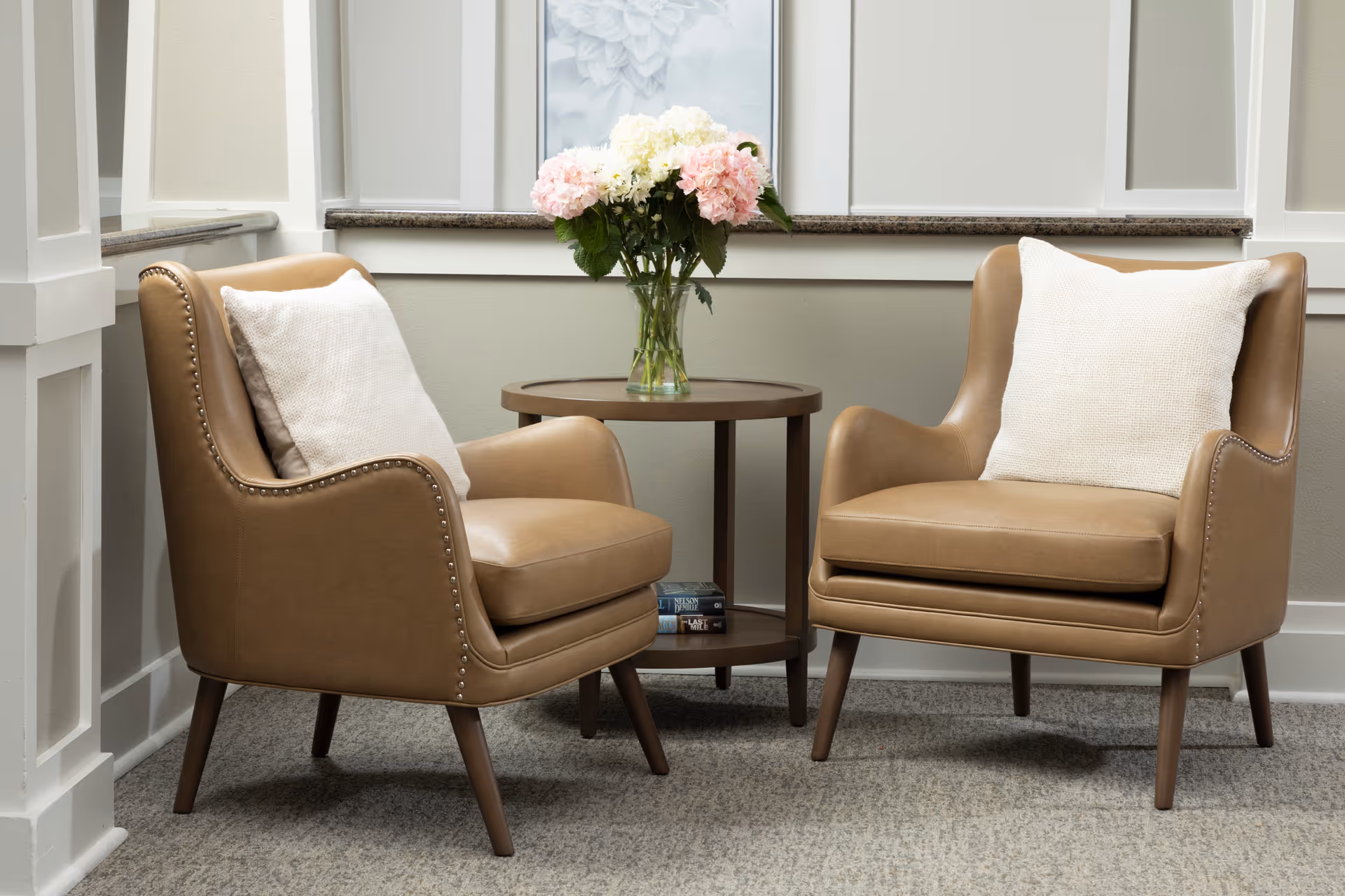 Two tan leather armchairs with white cushions are positioned facing each other with a round wooden side table between them. On the table is a glass vase filled with pink and white flowers. The setting is indoors with light-colored walls and carpeted floor.