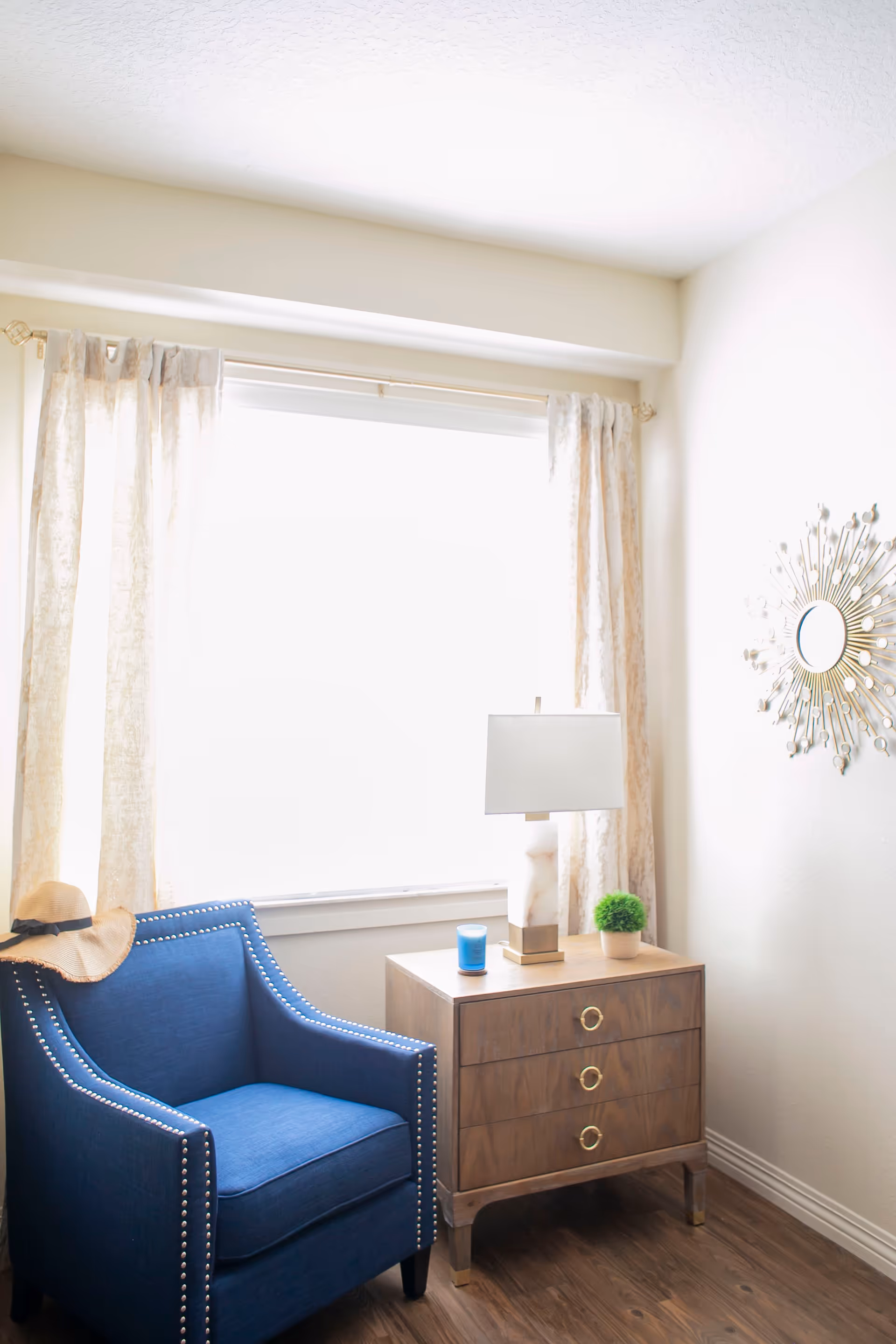 A cozy corner of a room featuring a blue armchair with silver studs, a wooden three-drawer nightstand with gold ring handles, a white and gold table lamp, a small green potted plant, a blue candle, and a decorative sunburst mirror on the wall. Light beige curtains frame a large window letting in bright natural light.