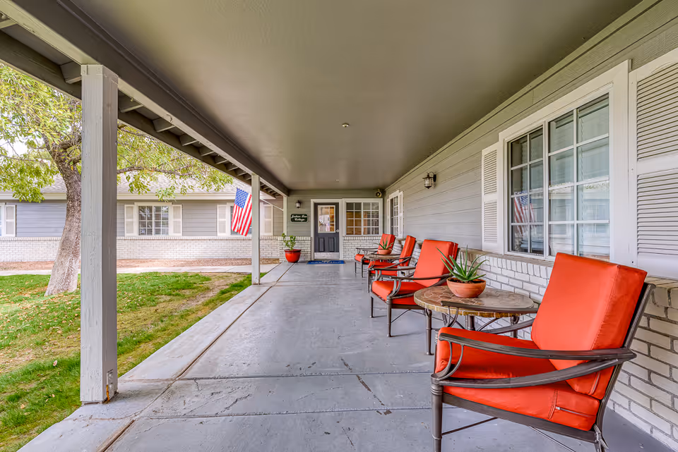 Covered front porch with red cushioned chairs and small tables leading to the entrance of a senior living building.