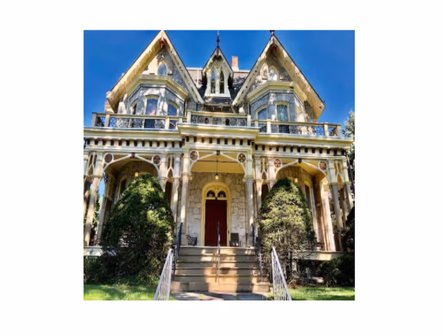 Front exterior view of a large, ornate Victorian-style house with a steeply pitched roof, decorative trim, and a central red door accessed by a set of stairs with metal railings. The house is surrounded by greenery and bushes under a clear blue sky.