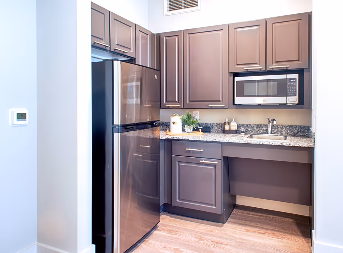 A modern kitchen area with dark brown cabinets, a stainless steel refrigerator, a built-in microwave, a small sink, and granite countertops. The floor is wood, and there is a small plant and some kitchen items on the counter.
