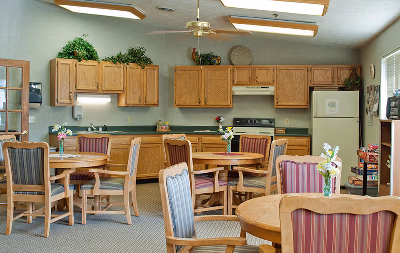 Dining area with round wooden tables and upholstered chairs in front of a kitchen with oak cabinets, refrigerator, and stove.