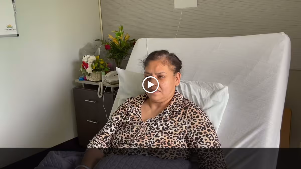 A woman wearing a leopard print top is sitting up in a bed with white sheets and a pillow behind her. Next to the bed is a nightstand with a telephone, a basket of flowers, and a vase with yellow and red flowers. The room has light-colored walls and a framed picture on the wall.