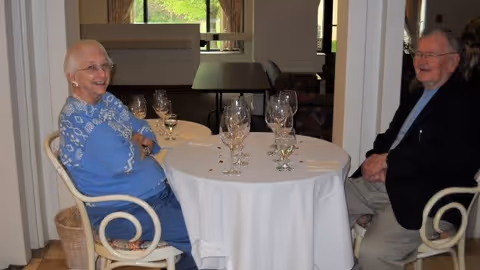 An elderly woman and an elderly man sitting at a round table covered with a white tablecloth in a dining area. The table has several empty wine glasses and some small decorative items. The woman is wearing a blue sweater and glasses, smiling, while the man is wearing a dark jacket and glasses, also smiling. The background shows windows with greenery outside.