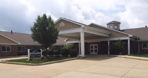 Exterior view of RidgeCrest Health Campus building with a covered entrance, a tree, and a sign displaying the facility's name. The sky is partly cloudy.