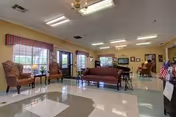 A spacious common area in a senior living facility with large windows allowing natural light. The room features patterned armchairs, a wooden bench, a wheelchair, and a table with chairs in the background. The floor has a tiled pattern, and the ceiling has fluorescent lighting fixtures.
