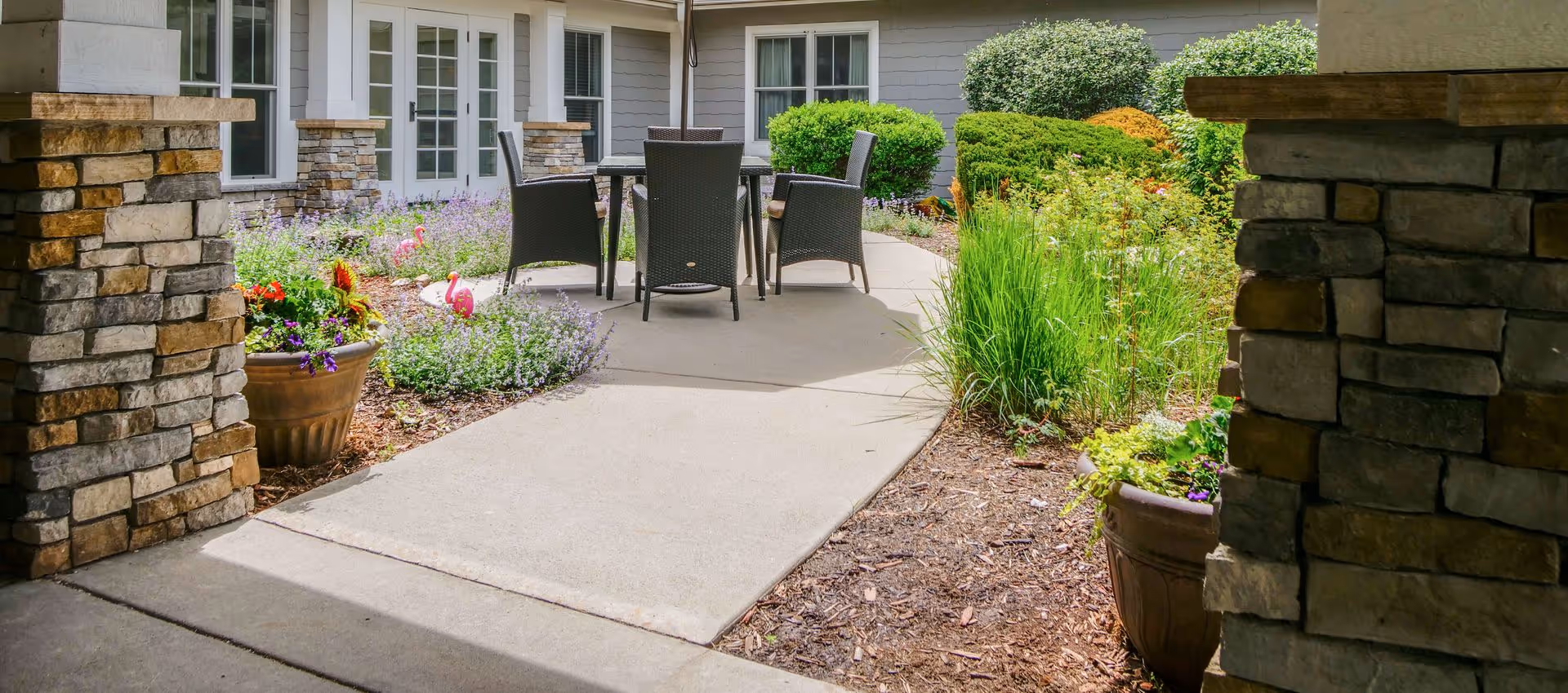 Outdoor patio area with a concrete walkway leading to a round table surrounded by four wicker chairs. The patio is bordered by flower beds with various green shrubs, flowering plants, and decorative pink flamingos. The building exterior features stone pillars and gray siding with white-framed windows and doors.