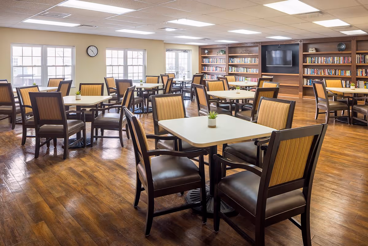 A bright and spacious dining room with multiple square tables, each surrounded by four chairs with striped upholstery. Small potted plants are placed on each table. The room features large windows letting in natural light, a clock on the wall, and a wooden bookshelf filled with books and a mounted flat-screen TV.