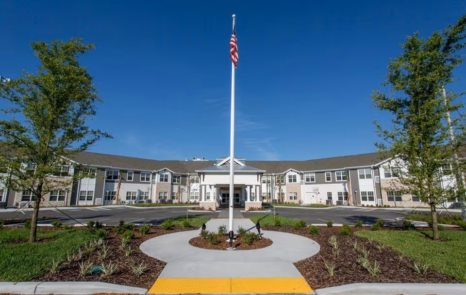 Front exterior view of The Crossings at Riverview senior living facility with a flagpole in the center, landscaped garden beds, and a clear blue sky.