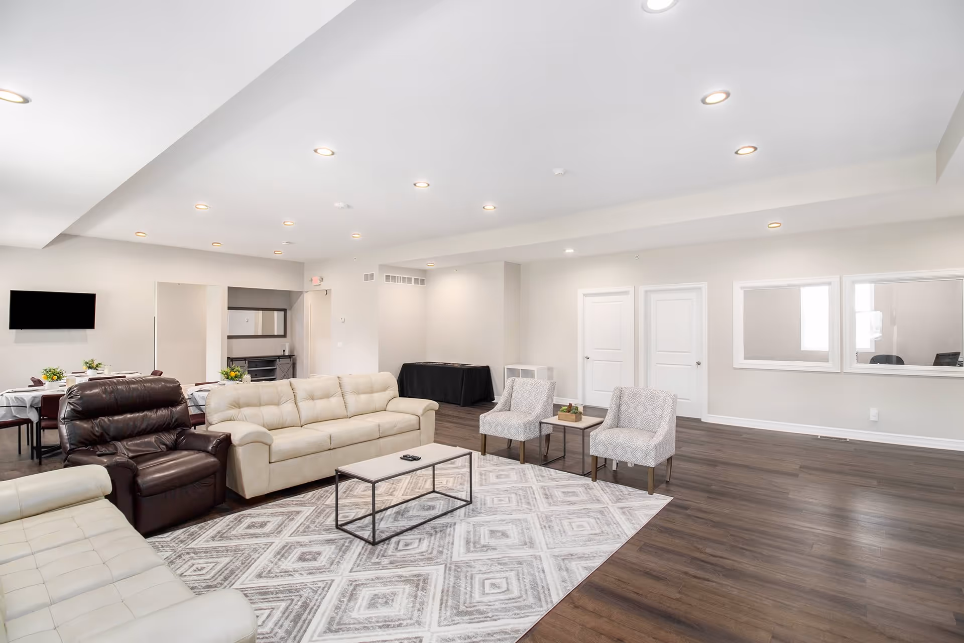 A spacious and well-lit living room area with beige and brown leather sofas, two patterned armchairs, a rectangular coffee table on a geometric patterned rug, and a wall-mounted flat-screen TV. The room has recessed ceiling lights, wood flooring, and light-colored walls with two white doors and two large windows on one side.