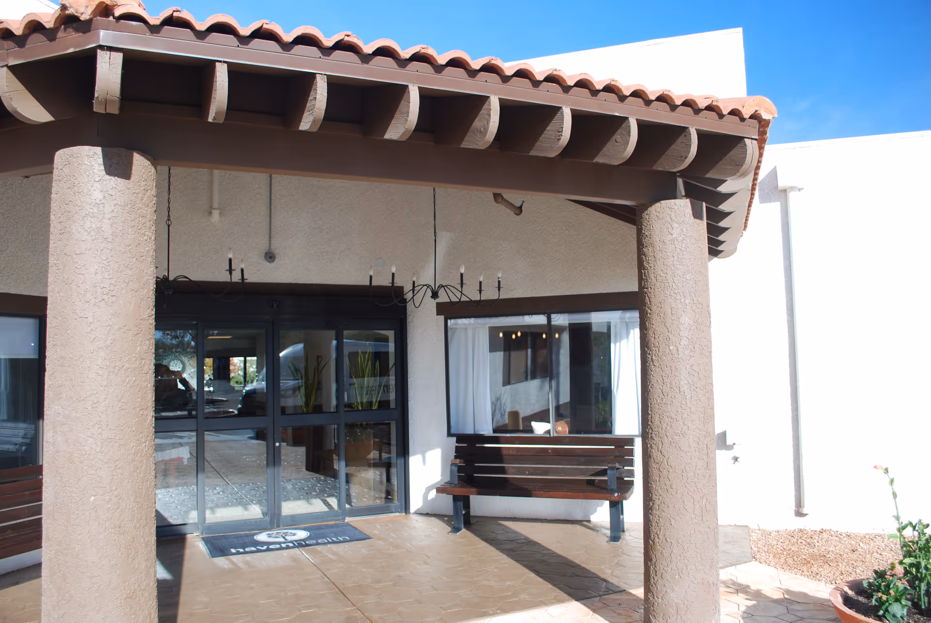 Covered entrance of a building with stucco columns, clay tile overhang, glass double doors, a bench, and a welcome mat.
