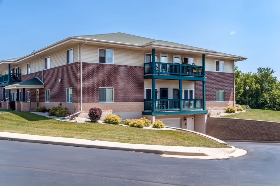 Exterior view of a two-story senior living facility building with brick and beige siding, green balconies, and a sloped driveway leading to a garage. The building is surrounded by well-maintained grass, shrubs, and trees under a clear blue sky.