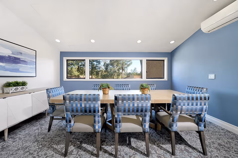 A modern dining room with a rectangular wooden table surrounded by eight blue and gray patterned chairs. The room has a blue accent wall with three rectangular windows showing trees outside. There is a white sideboard with a small green plant and a blue abstract painting on the adjacent white wall. The floor is covered with a patterned carpet, and recessed ceiling lights illuminate the space.