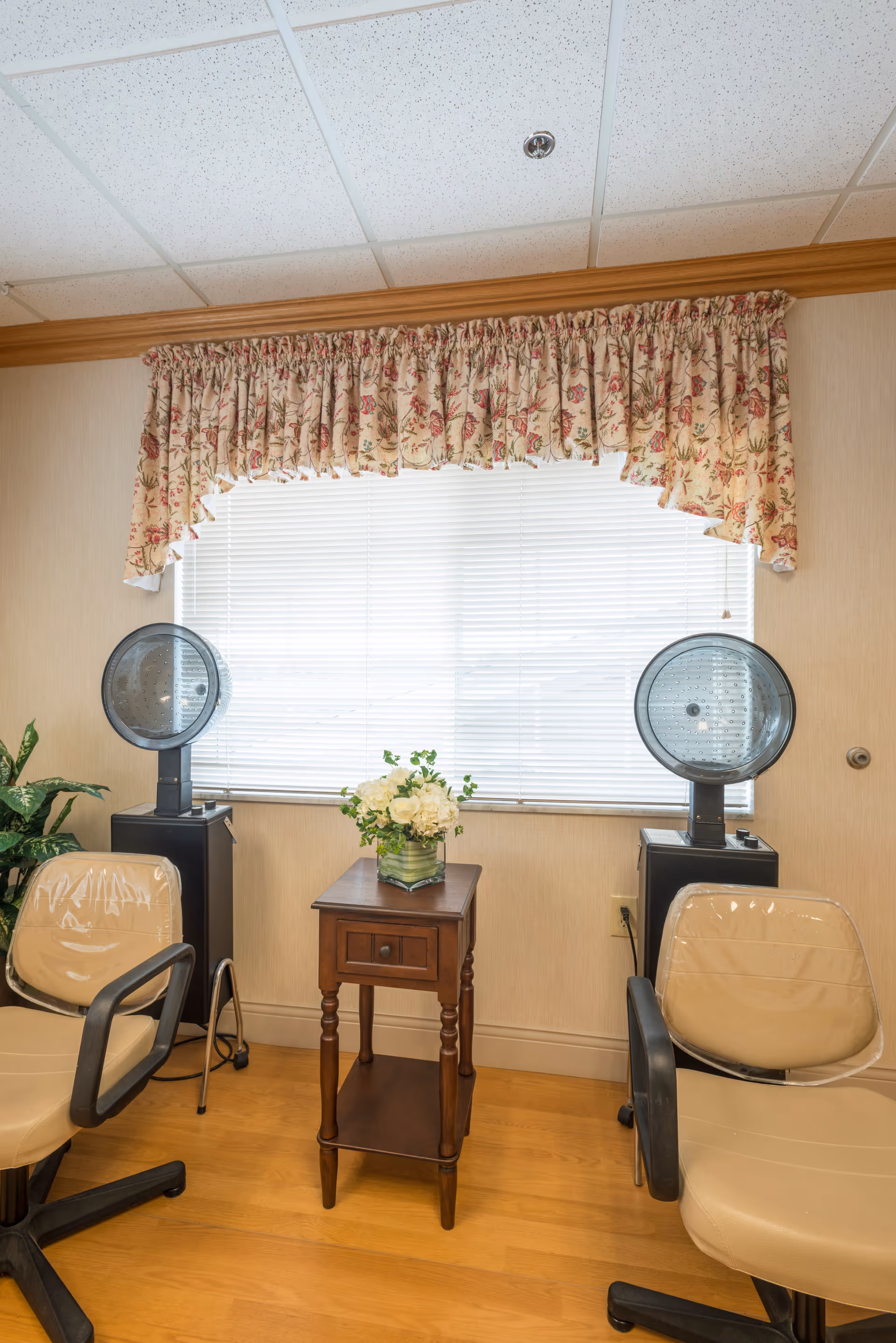 Interior view of a room with two beige salon chairs covered in plastic, two hair dryer machines on stands, a small wooden table with a drawer and a vase of white flowers, and a window with closed blinds and floral valance curtains.
