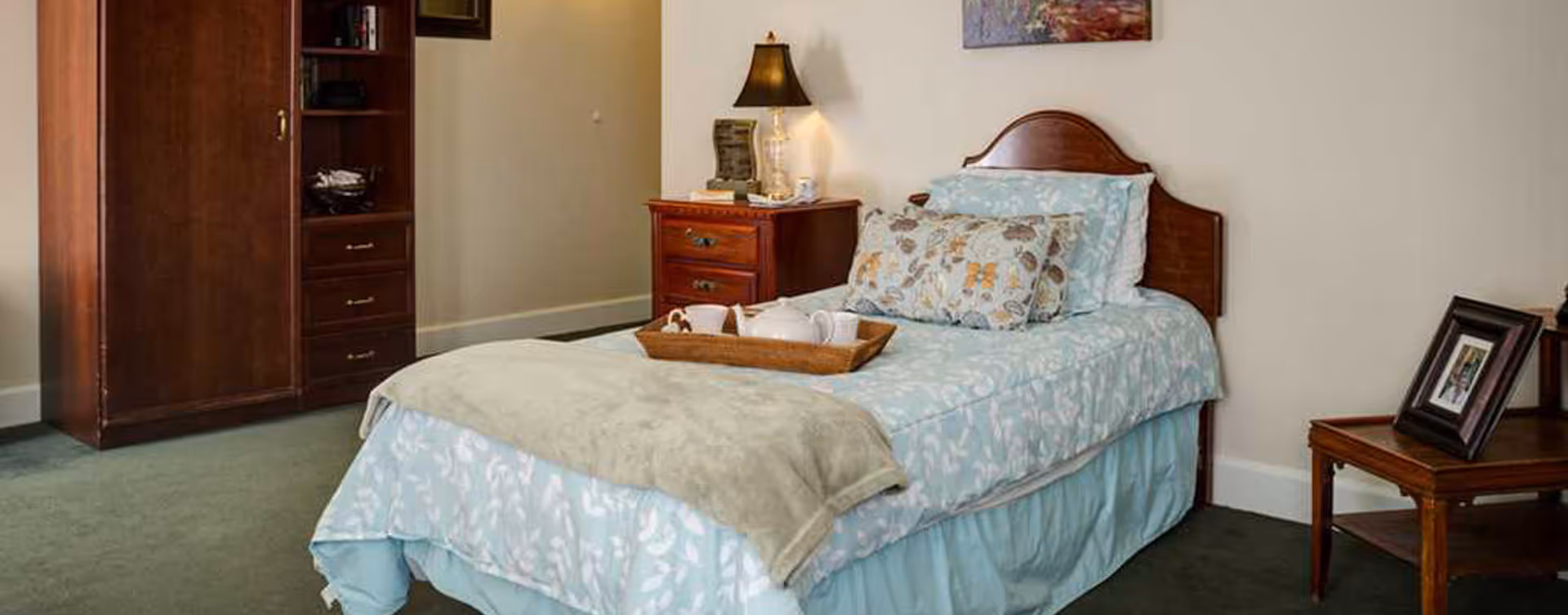A neatly made single bed with light blue floral bedding and a beige blanket at the foot. A wooden tray with a teapot and cups sits on the bed. To the right of the bed is a wooden nightstand with a lamp and decorative items. There is a wooden cabinet with drawers and shelves against the wall on the left side. A small wooden table with a framed photo is on the right side of the image. The room has green carpet and light-colored walls with a painting hanging above the bed.