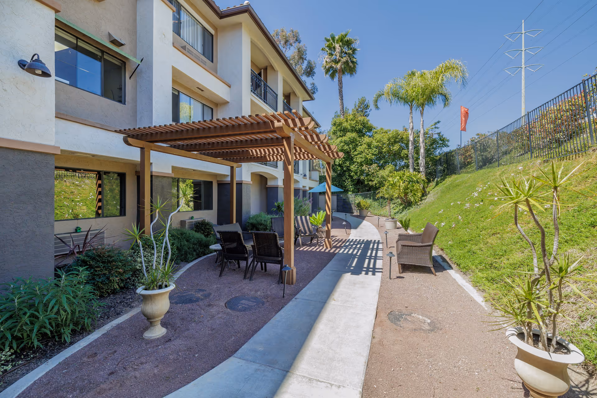 Outdoor patio area at Rancho Penasquitos Senior Living featuring a wooden pergola with several chairs underneath, potted plants, a concrete pathway, and a grassy hill with palm trees and other greenery under a clear blue sky.