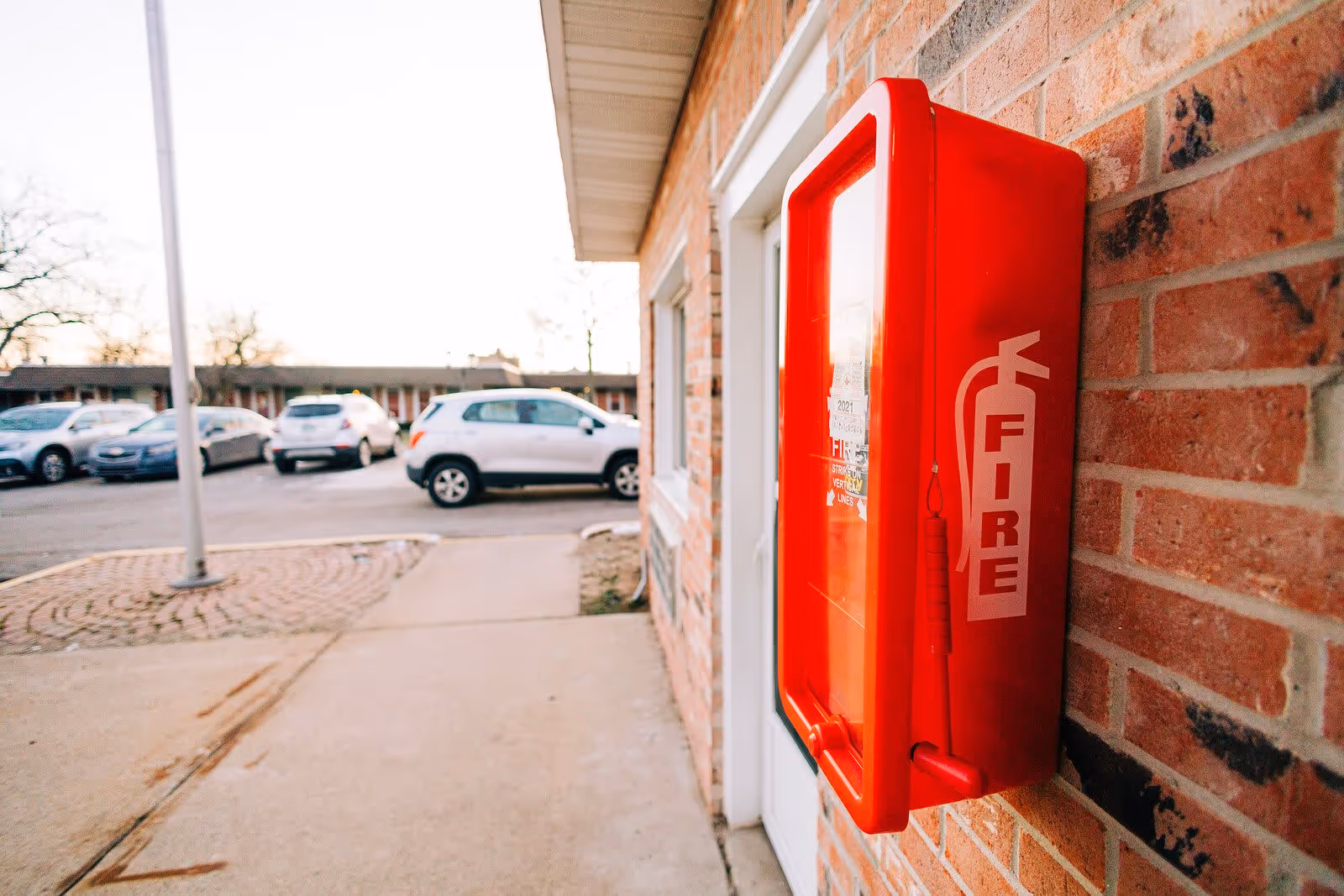 A red fire extinguisher box mounted on a brick wall outside a building, with a parking lot and several parked cars visible in the background.