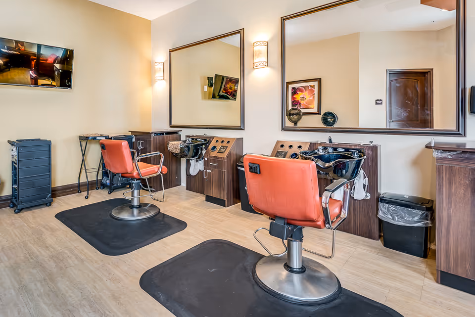 Interior view of a salon area with two red salon chairs facing large mirrors mounted on the wall. Each station has a black wash basin and wooden cabinetry. The room has light-colored walls and flooring, with a wall-mounted TV and a small trash bin visible.