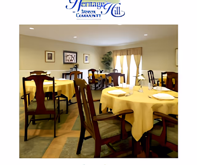 A dining room with several round tables covered with yellow tablecloths, each set with plates and napkins. Wooden chairs with cushioned seats surround the tables. The room has beige walls, framed pictures, a large window with curtains, and a potted plant in the corner.