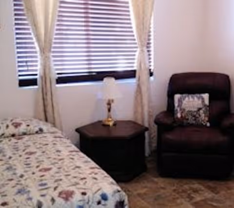 A cozy bedroom with a single bed covered in a floral patterned bedspread, a dark wooden nightstand with a small lamp, and a dark upholstered armchair with a decorative pillow. The window behind has closed blinds and light-colored curtains.