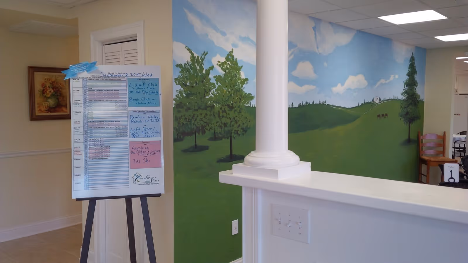 Interior view of a senior living facility with a painted mural of trees and a blue sky on the wall. A white column and half wall are in the foreground, and a schedule board on an easel lists daily activities. There is a wooden chair and a table with a number 1 sign in the background.