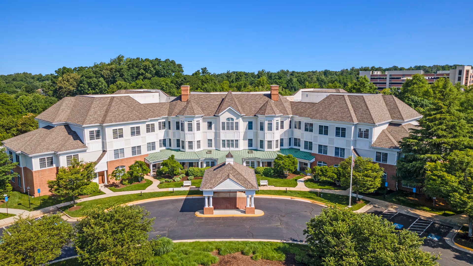 Aerial view of a large, multi-story senior living facility building with a circular driveway and a covered entrance. The building has a combination of white siding and red brick exterior with multiple windows and a brown shingled roof. Surrounding the building are well-maintained green lawns, trees, and shrubs under a clear blue sky.