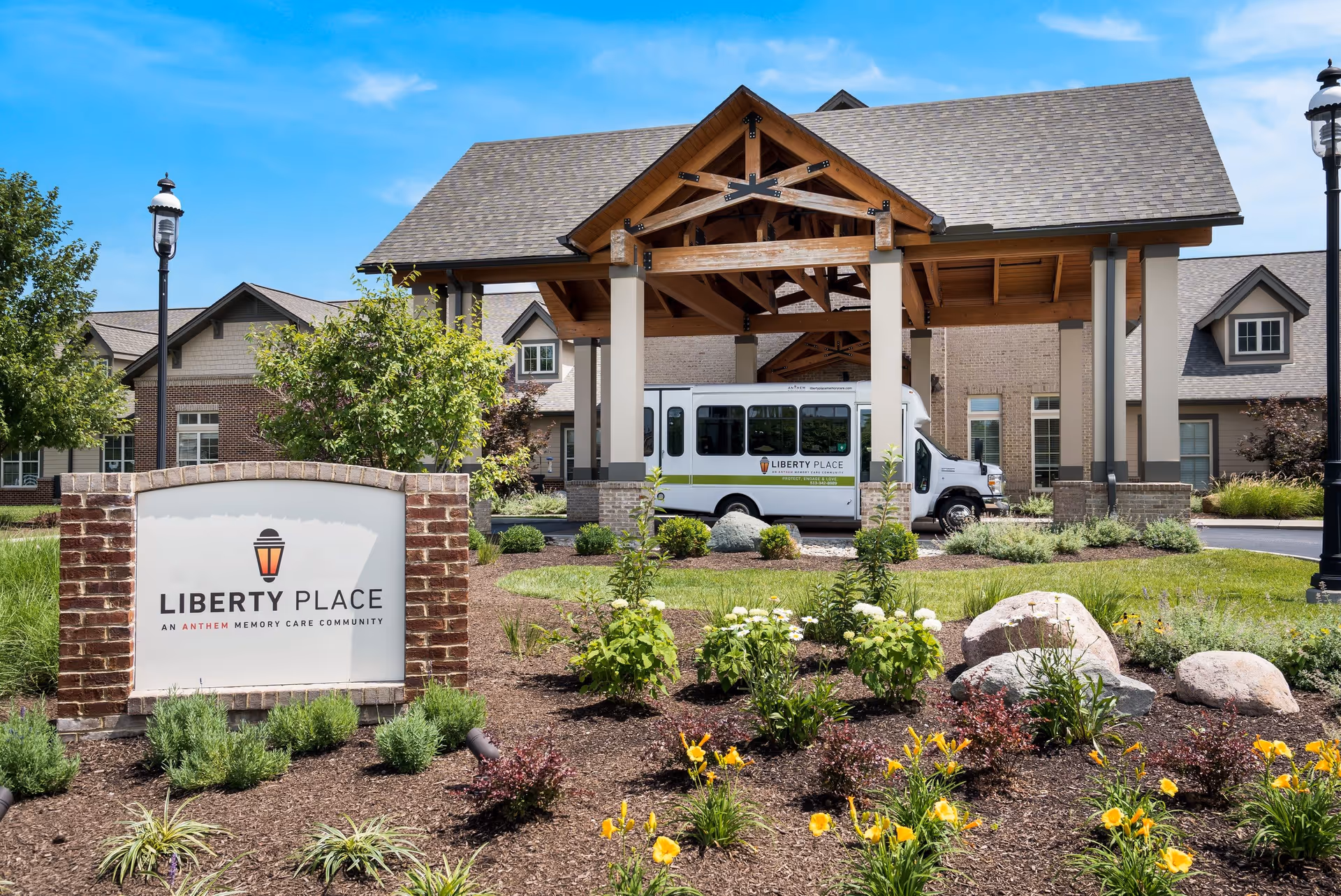 Exterior view of Liberty Place Memory Care facility showing a landscaped garden with flowers and shrubs in the foreground, a brick sign with the facility name, a covered entrance with wooden beams, and a white Liberty Place shuttle bus parked under the entrance canopy.
