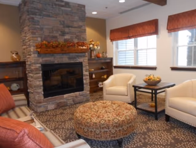 Cozy communal living room with a stone fireplace, armchairs, a patterned ottoman and a side table beneath windows.