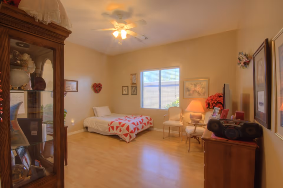 Well-lit assisted living bedroom with a single bed topped by a red-and-white quilt, chairs, a lamp, dresser and display cabinet.
