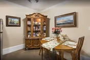 A dining room with a wooden dining table set for four, featuring a floral table runner and a vase with pink flowers in the center. Behind the table is a wooden china cabinet displaying various decorative items and dishes. The walls are adorned with two framed paintings, and the room has a beige carpet and light-colored walls.