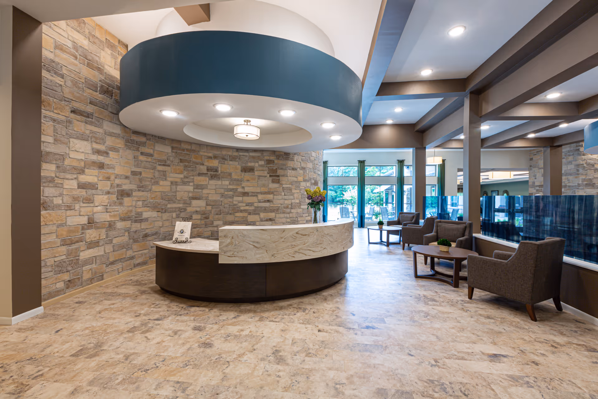 A modern senior living facility lobby with a curved reception desk in front of a stone accent wall. The ceiling above the desk features a circular design with recessed lighting and a central light fixture. To the right, there is a seating area with several armchairs and coffee tables, and large windows in the background let in natural light and provide a view of an outdoor patio area.