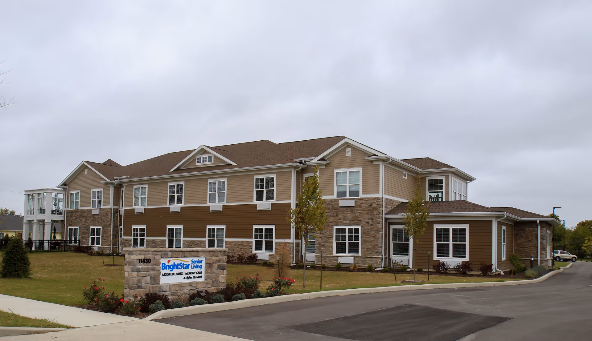Exterior view of a two-story senior living facility building with beige and brown siding and stone accents. There is a landscaped lawn with small trees and bushes, and a stone sign in front that reads 'BrightStar Senior Living Assisted Living | Memory Care A Higher Standard'. The sky is overcast.