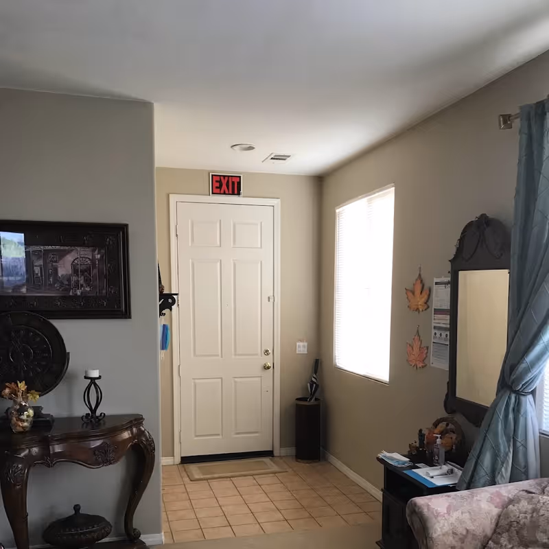 Interior view of a room with a white exit door, a window with blinds, a decorative mirror on the wall, a small table with various items, and a wooden console table with a candle and decorative items. The walls are painted beige and there is a tiled floor near the door.