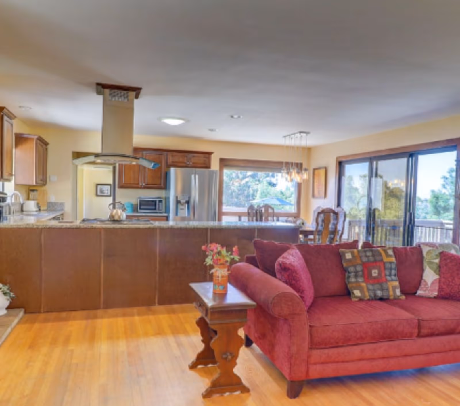 Open-concept living area with a red sofa in the foreground, a kitchen island and stainless-steel refrigerator in the background, and sliding glass doors to the right.
