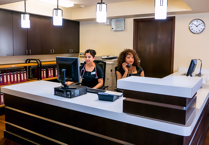 Reception area with two women behind a large white and dark wood desk. One woman is using a computer while the other is speaking on a phone. The background shows dark wood cabinets, shelves with red binders, a clock on the wall, and a door.