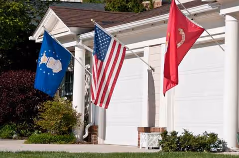 Front of a white building with three flags by columns at Falcons Landing West Falls Center