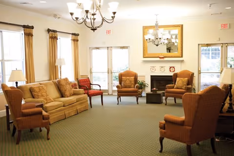 A bright and spacious senior living common area with beige and brown upholstered chairs and a sofa arranged around a central coffee table. The room features large windows with curtains, two chandeliers, a fireplace with a large mirror above it, and exit doors leading outside.