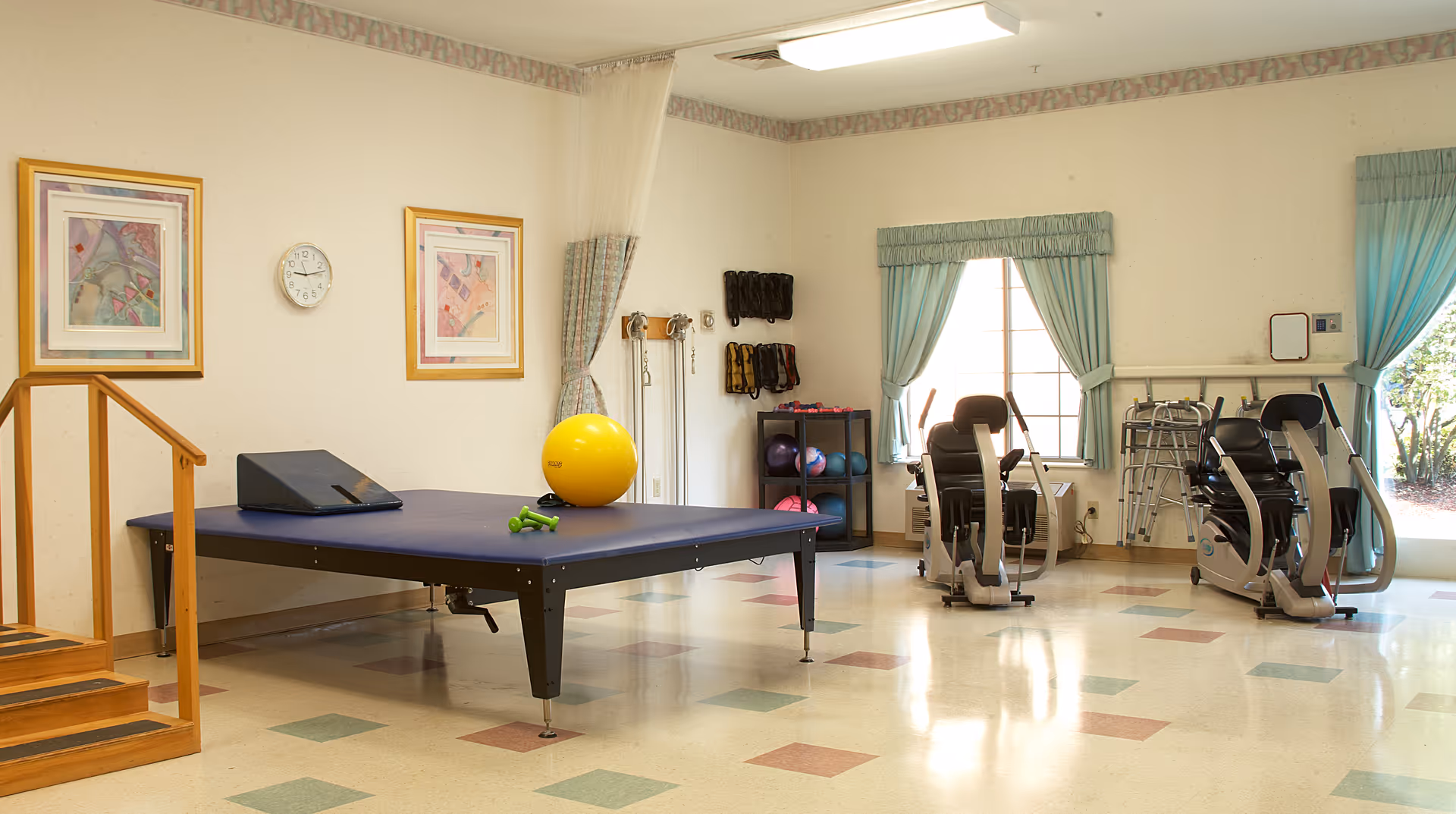 A physical therapy or exercise room with a large padded table, a yellow exercise ball, small green dumbbells, two exercise machines, a wooden staircase for therapy, and various exercise equipment on shelves and hanging on the wall. The room has light-colored walls, two framed pictures, a clock, and windows with green curtains letting in natural light.