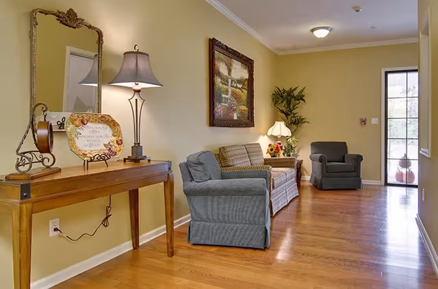 A cozy living room area with wooden flooring, featuring a wooden console table with a decorative mirror and lamp on the left. There are two upholstered armchairs and a sofa along the right wall, with a framed painting and a potted plant in the corner. A door with glass panels is visible at the far end of the room.