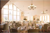 A spacious dining room with large windows allowing natural light to fill the space. The room is furnished with round tables covered with white tablecloths, set with plates, glasses, and napkins. Comfortable chairs surround each table, and elegant chandeliers hang from the ceiling.