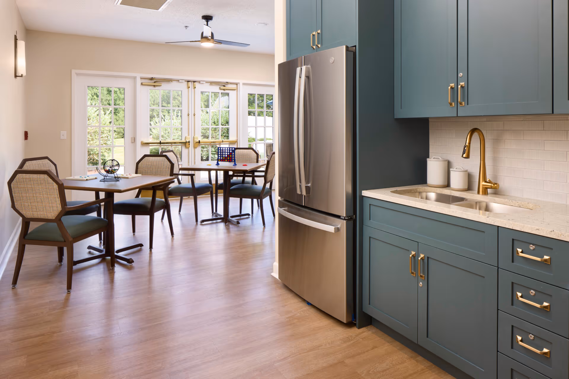A bright room featuring a modern kitchen area with teal cabinets, a stainless steel refrigerator, and a gold faucet over a double sink. Adjacent to the kitchen are two tables with chairs, each set with a game, and large glass doors leading outside, allowing natural light to fill the space.