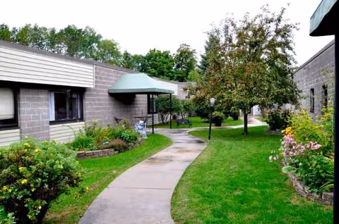 Grassy courtyard with a winding concrete path, trees, flowering beds, and an umbrella-covered seating area.