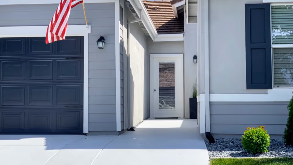 Front entrance of a house with a gray garage door on the left, an American flag mounted above the garage, a white door with glass panels in the center, and a window with black shutters on the right. There are small bushes and decorative rocks near the entrance.
