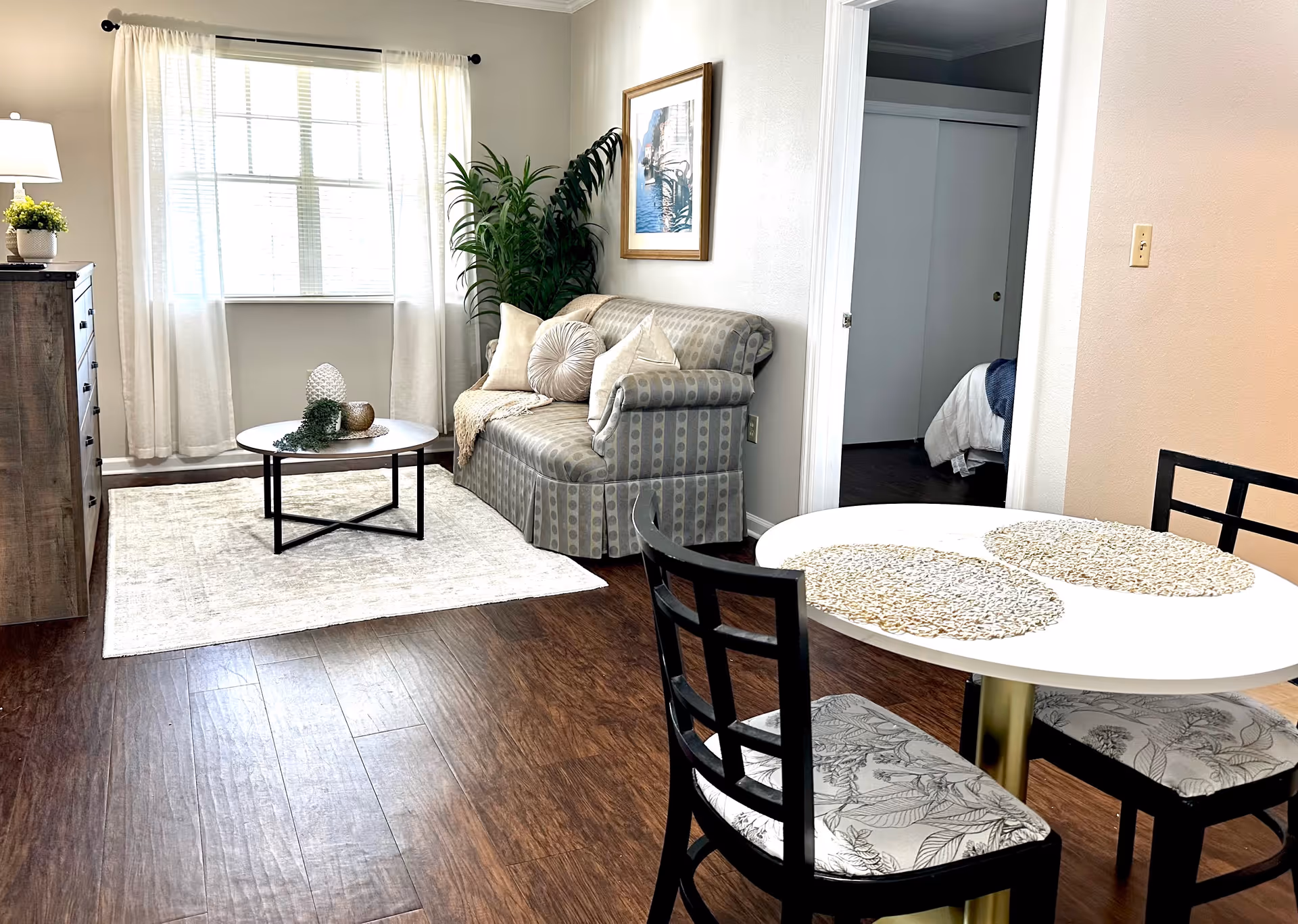 Bright furnished living area with a small sofa, coffee table on a rug, dining table with chairs, dresser and a doorway to a bedroom.