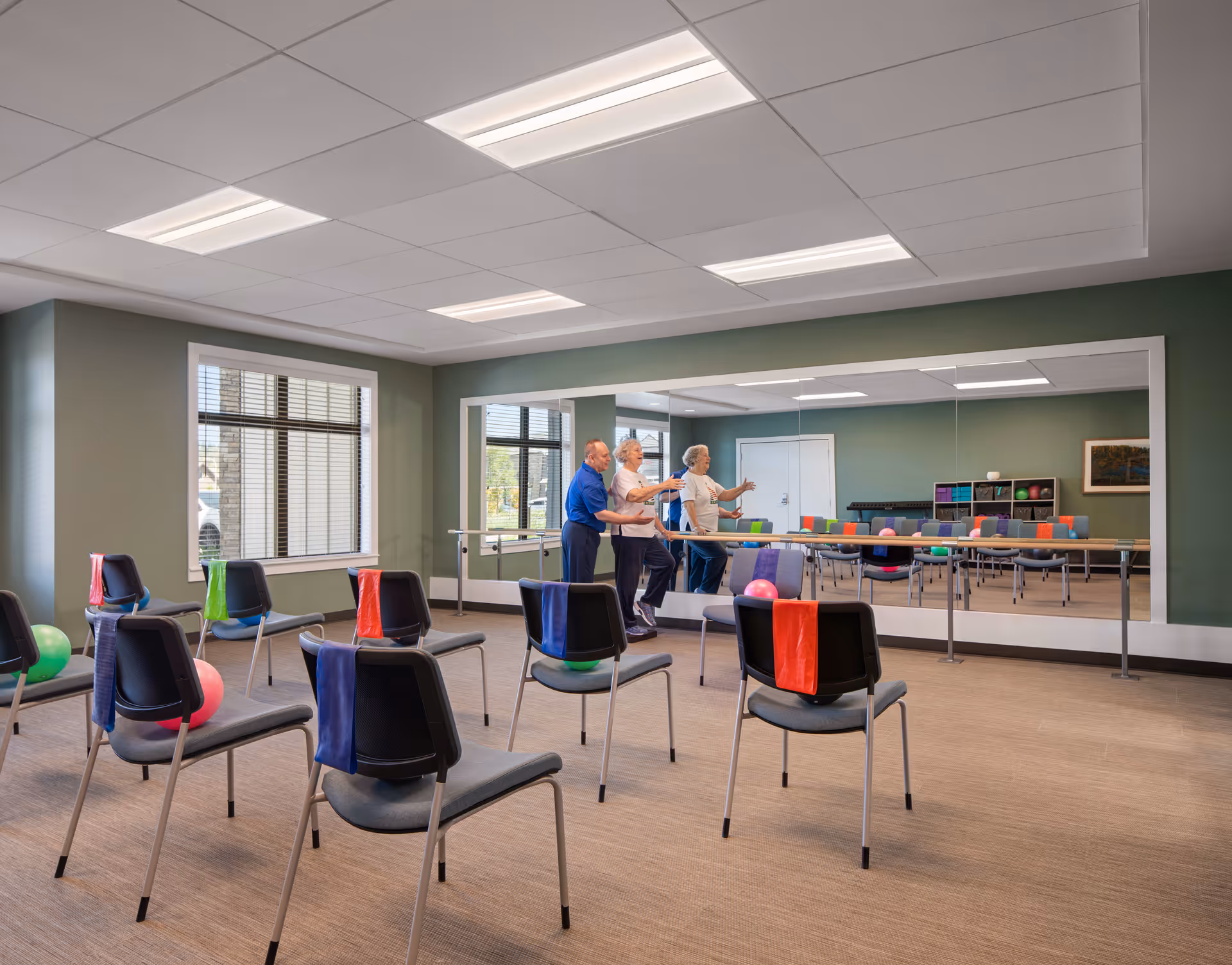 A fitness or activity room with chairs spaced apart, each with a colorful resistance band draped over the back. Three elderly individuals are standing near a ballet barre in front of a large mirror, engaging in a group exercise. The room has green walls, large windows, and bright overhead lighting.